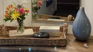 Une pièce comprenant une commode en bois, un miroir, un lustre, un vase avec des fleurs et un plateau en osier avec un savonnière.