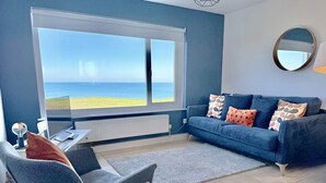 Un salon moderne avec une grande fenêtre donnant sur la plage, un canapé bleu orné de coussins à motifs, un fauteuil gris et un miroir rond accroché au mur.