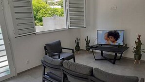 A modern living room with a sofa, a coffee table, and a TV. There are plants and a window with shutters.