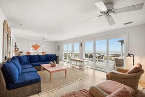 Un salon spacieux avec un grand canapé d’angle, une petite table et un fauteuil en osier. Il y a un ventilateur de plafond et, grâce à de grandes portes coulissantes, on peut admirer la vue sur l’océan.