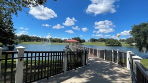 A pathway leading to a gazebo over a lake with a fountain, surrounded by trees and a clear sky.