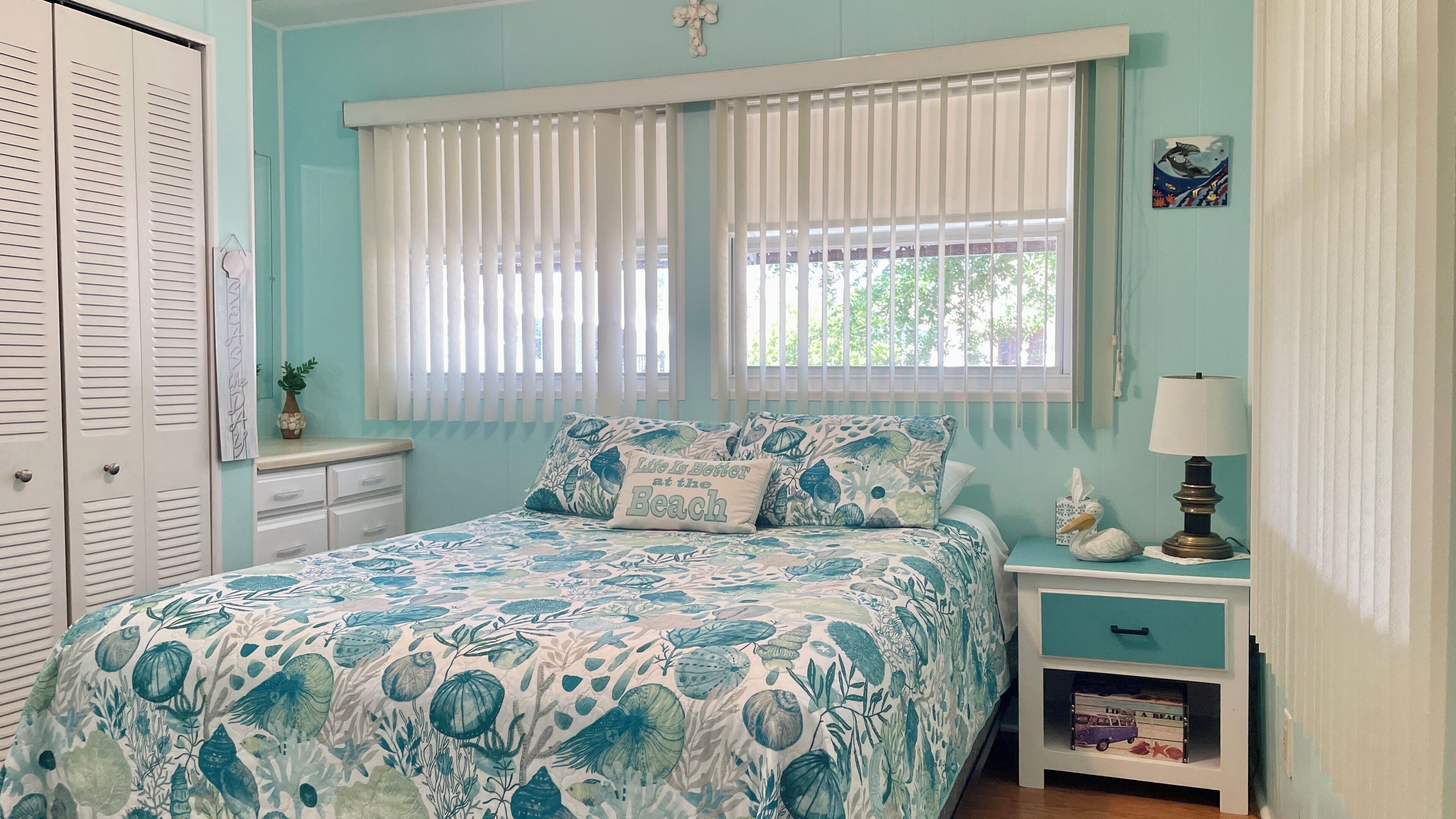 A bedroom with a bed, a nightstand, a lamp, a window with blinds, a wall decoration, and a door with louvered shutters.