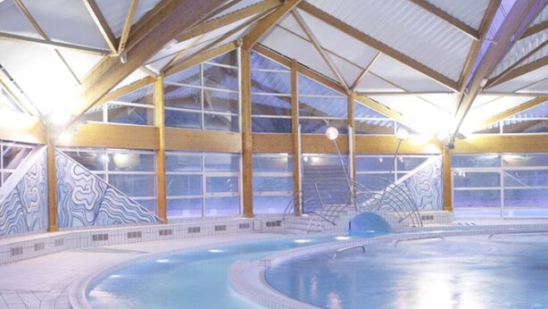 An indoor swimming pool with a curved design, large windows, and a high ceiling with wooden beams. - Studio Forêt (Amnéville)