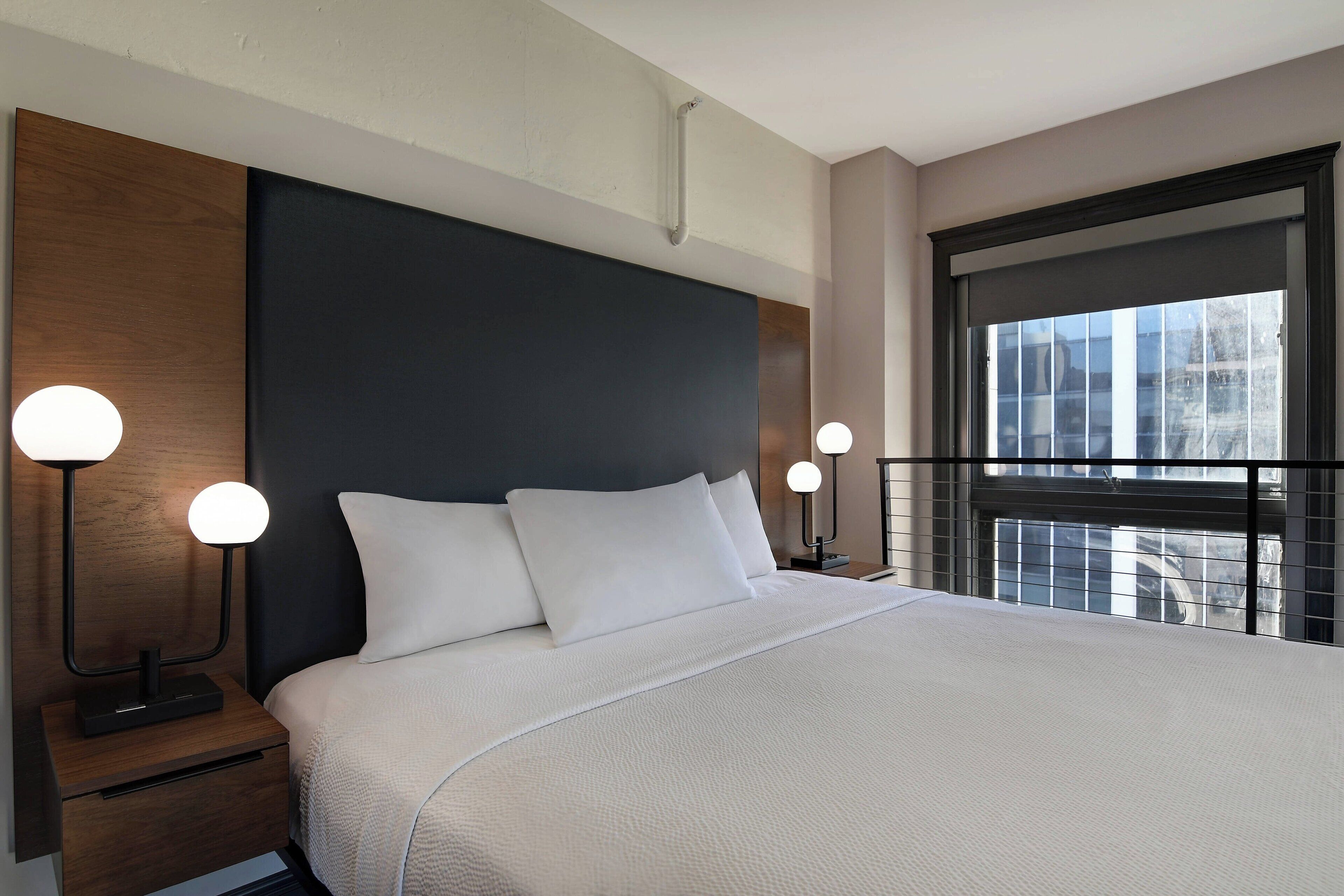 A modern hotel room with a large bed, two bedside tables with lamps, a window with a view, and a dark headboard.