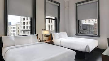 Habitación de hotel con dos camas, una mesita de noche, una lámpara y ventanas amplias con vistas a la ciudad.