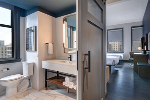 A modern hotel bathroom with a toilet, sink, and mirror. There are two beds in the adjacent room. - Courtyard by Marriott Cincinnati Downtown (Cincinnati)