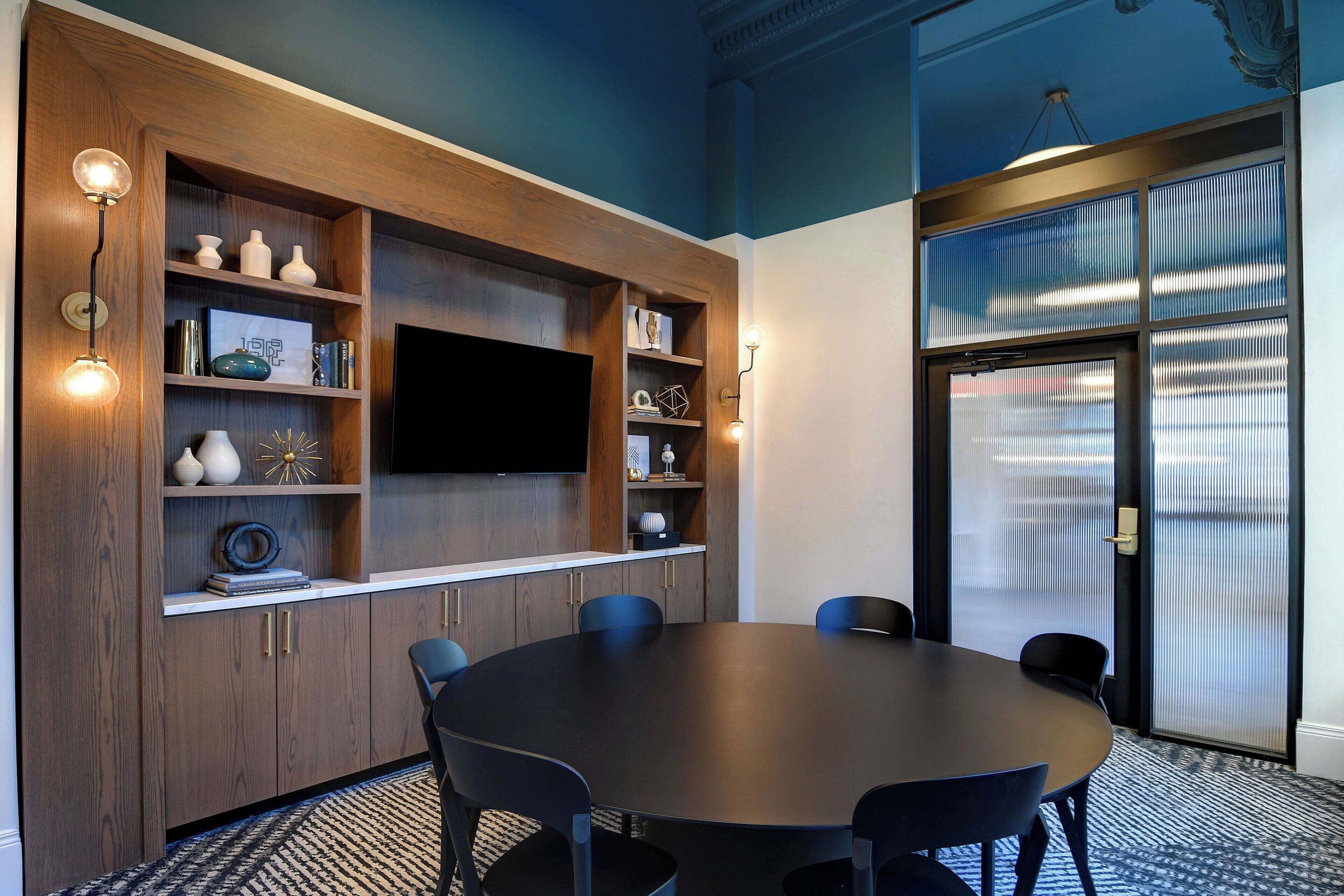 A conference room with a round table, chairs, a flat-screen TV, wooden shelving with decorative items, and a patterned carpet.
