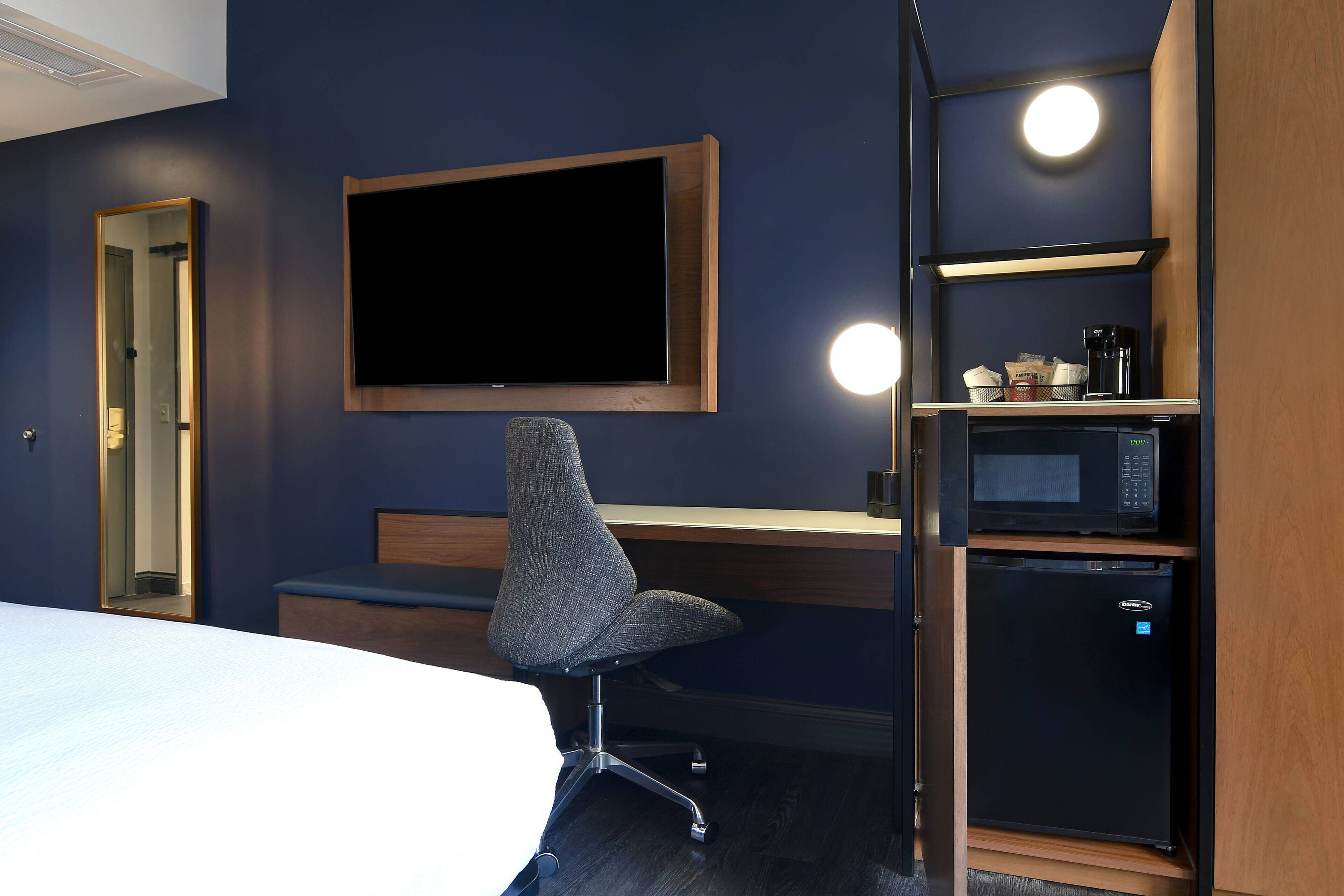 Habitación de hotel con cama, escritorio con silla, televisor de pantalla plana, cafetera, microondas y refrigerador.