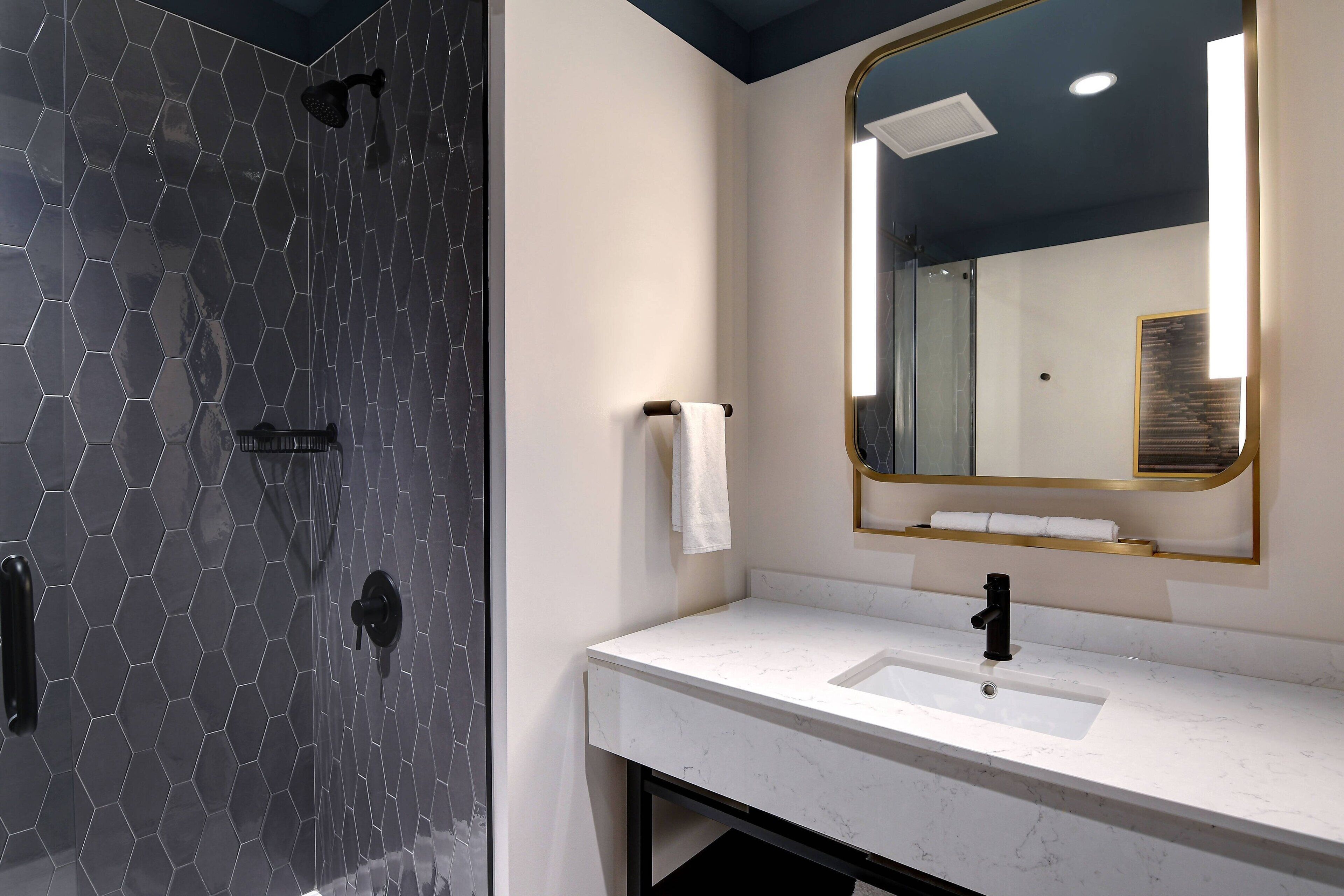 Un bagno moderno con doccia rivestita in piastrelle esagonali, lavandino bianco con rubinetto nero e un ampio specchio con cornice dorata.