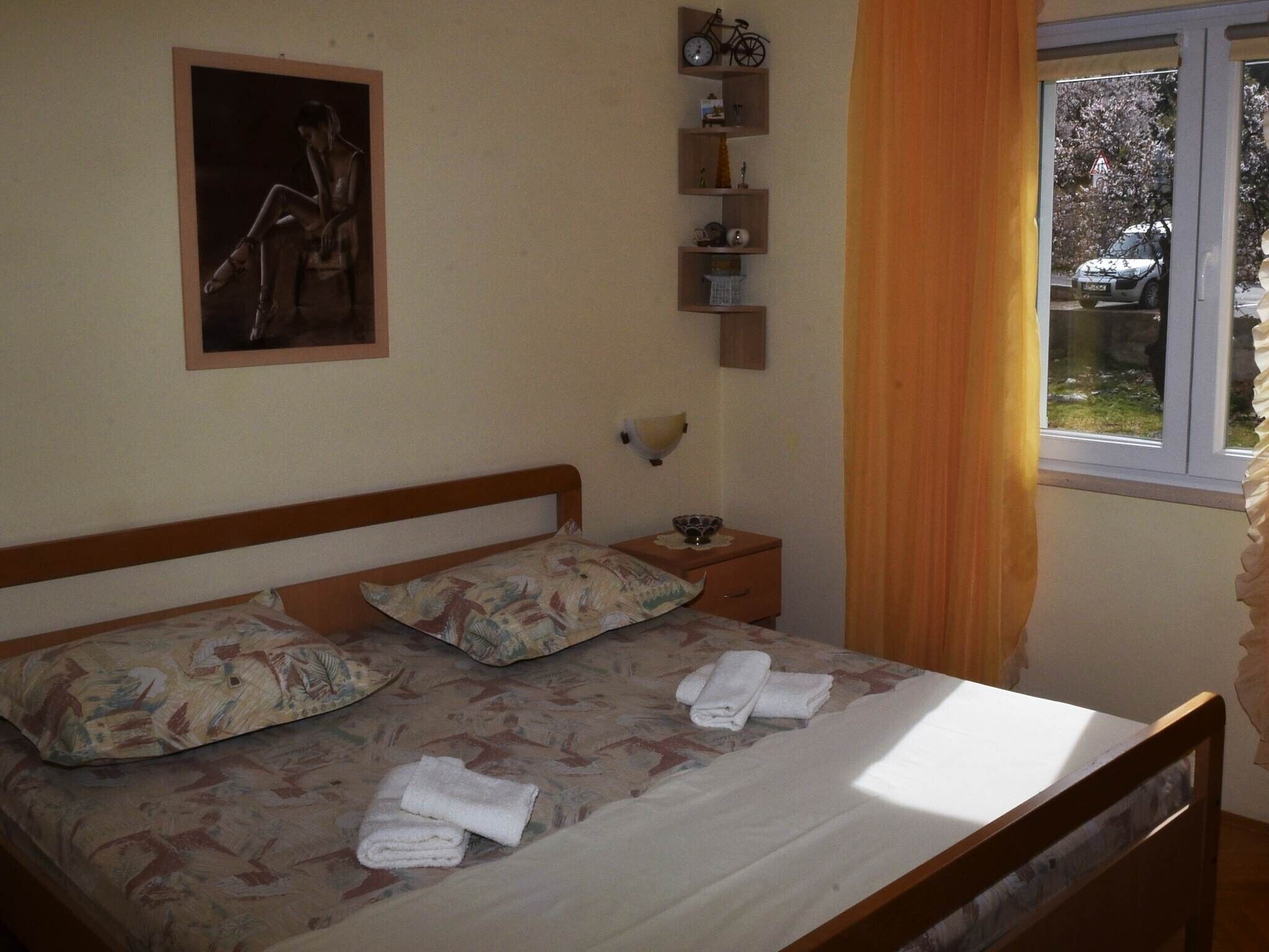 Une chambre à coucher avec un lit, des oreillers, une table de chevet, une étagère fixée au mur, une fenêtre avec des rideaux et un tableau accroché au mur.