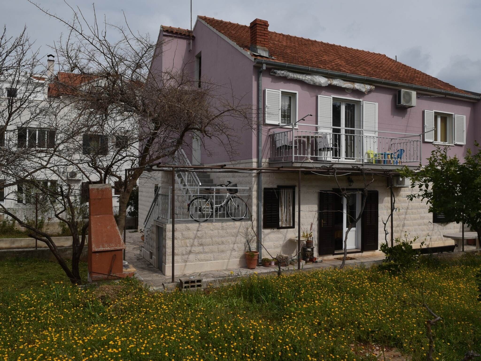 Una casa de dos pisos con un balcón, una escalera que lleva al nivel superior y un jardín con flores amarillas.