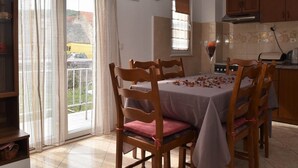 A dining area with a table set for six, wooden chairs, a kitchen area with cabinets and appliances, and a view through a sliding glass door to the outside.