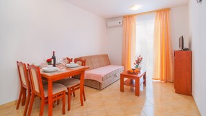 Un comedor con una mesa de madera, sillas y un sofá. Hay una mesita con fruta y una botella. Un equipo de aire acondicionado está montado en la pared.