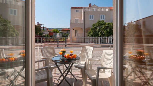 A balcony with a table set for breakfast, including fruits and drinks, overlooking a residential area with buildings and a tree. - Apartments Island Beat - Two Bedroom Apartment with Balcony (A9) (Supetar)
