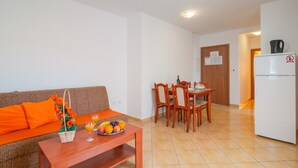 A living room with a sofa, a wooden table with fruit, and a dining area with chairs and a table. - Apartments Island Beat - Two Bedroom Apartment with Balcony (A9) (Supetar)
