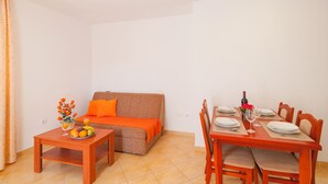 A dining area with a wooden table set for four, a sofa with orange cushions, and a fruit basket on a side table. - Apartments Island Beat - Two Bedroom Apartment with Balcony (A9) (Supetar)