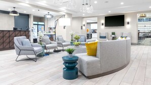 A modern hotel lobby with a reception desk, seating area with sofas and chairs, a coffee machine, and a TV screen.