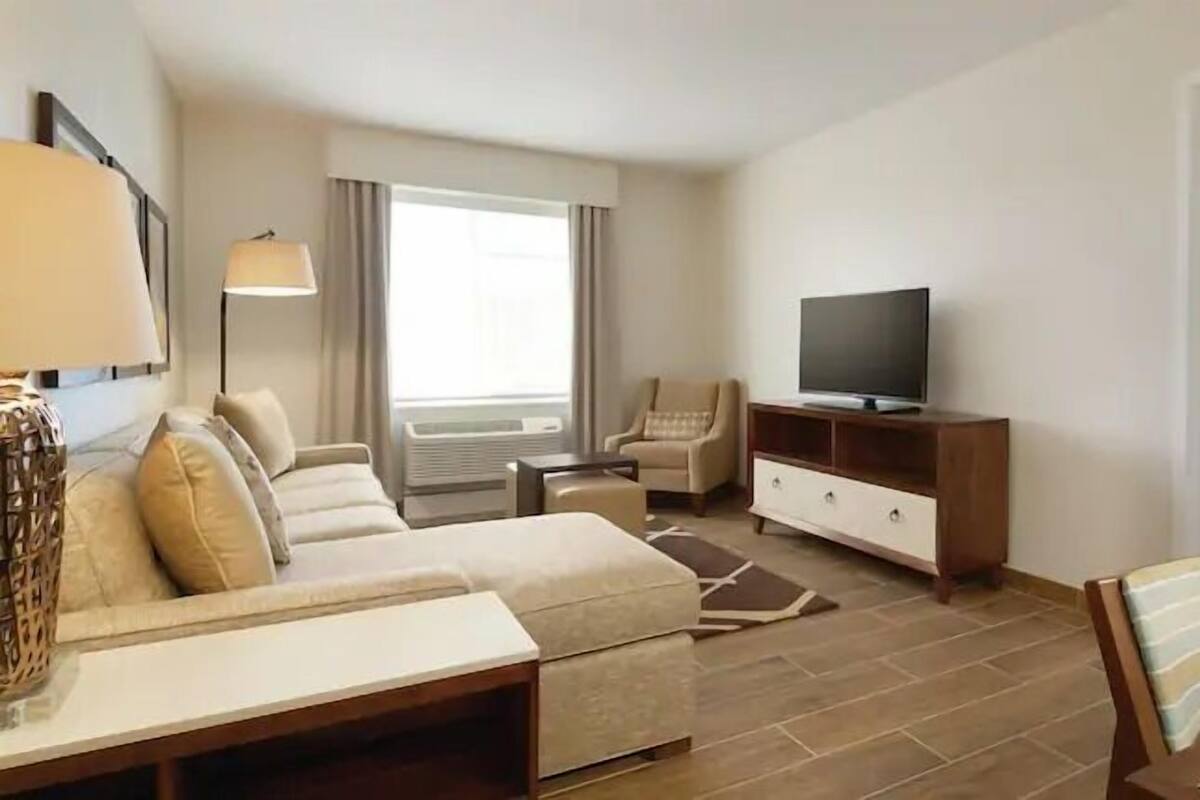 A hotel room with a sofa set, a TV on a wooden cabinet, a chair, and a bed with white linens.