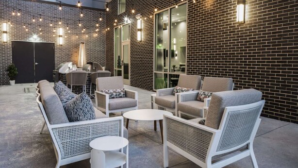 An outdoor seating area with wicker furniture and a grill, set against a brick wall with large windows.