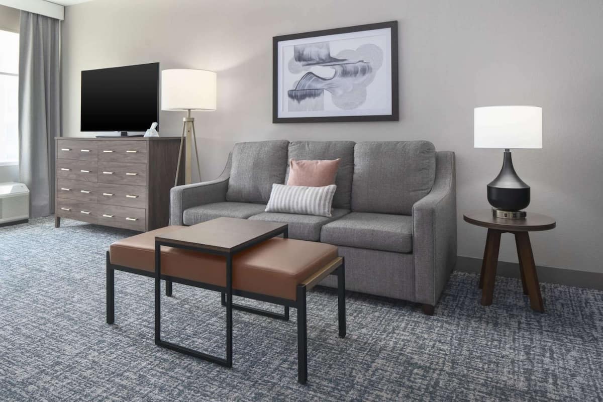 A modern hotel room with a grey sofa, a wooden side table, a flat-screen TV, a framed artwork, and a lamp.