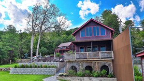 A two-story house with a balcony, a stone retaining wall, and a well-maintained lawn. - Lakeview Suites (Bolton Landing)