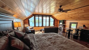 A bedroom with a large bed, a wooden ceiling, a fireplace, and a view of the outdoors. - Lakeview Suites (Bolton Landing)