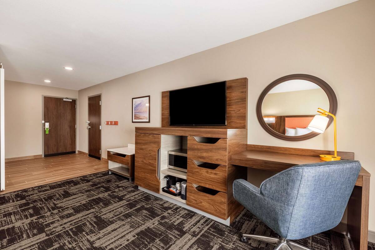 A hotel room with a wooden desk, a flat-screen TV, a mirror, and a blue chair.