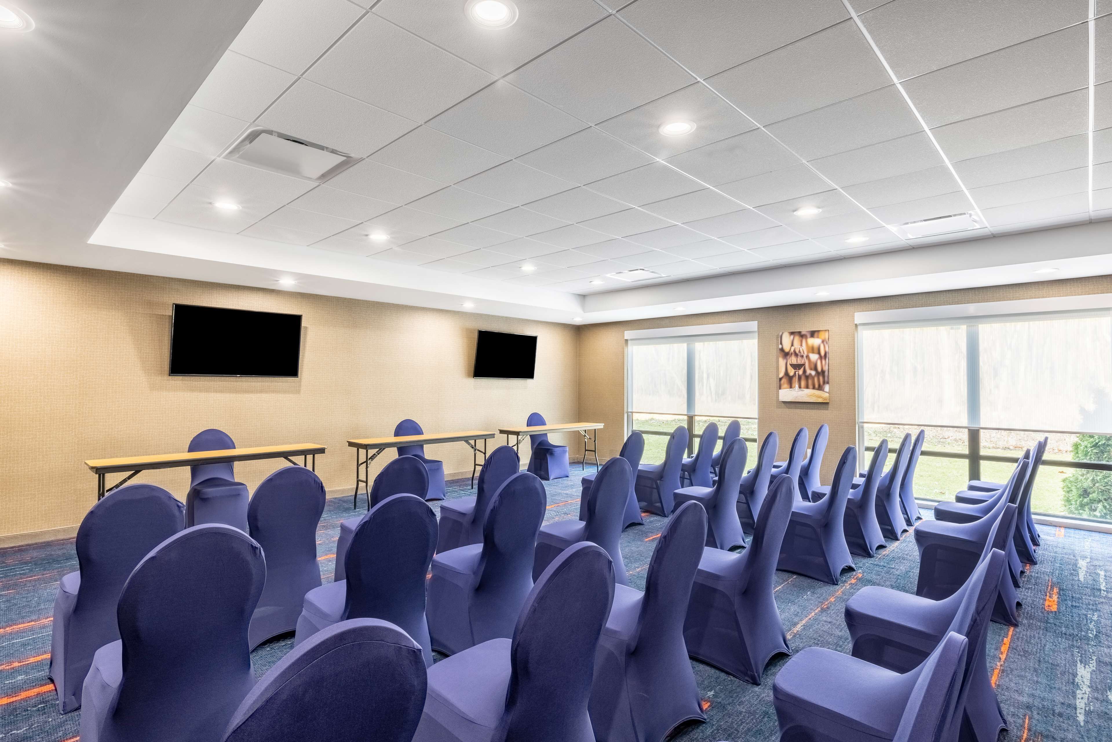 Un salón de conferencias con filas de sillas, una pantalla de proyector, mesas y ventanas amplias.