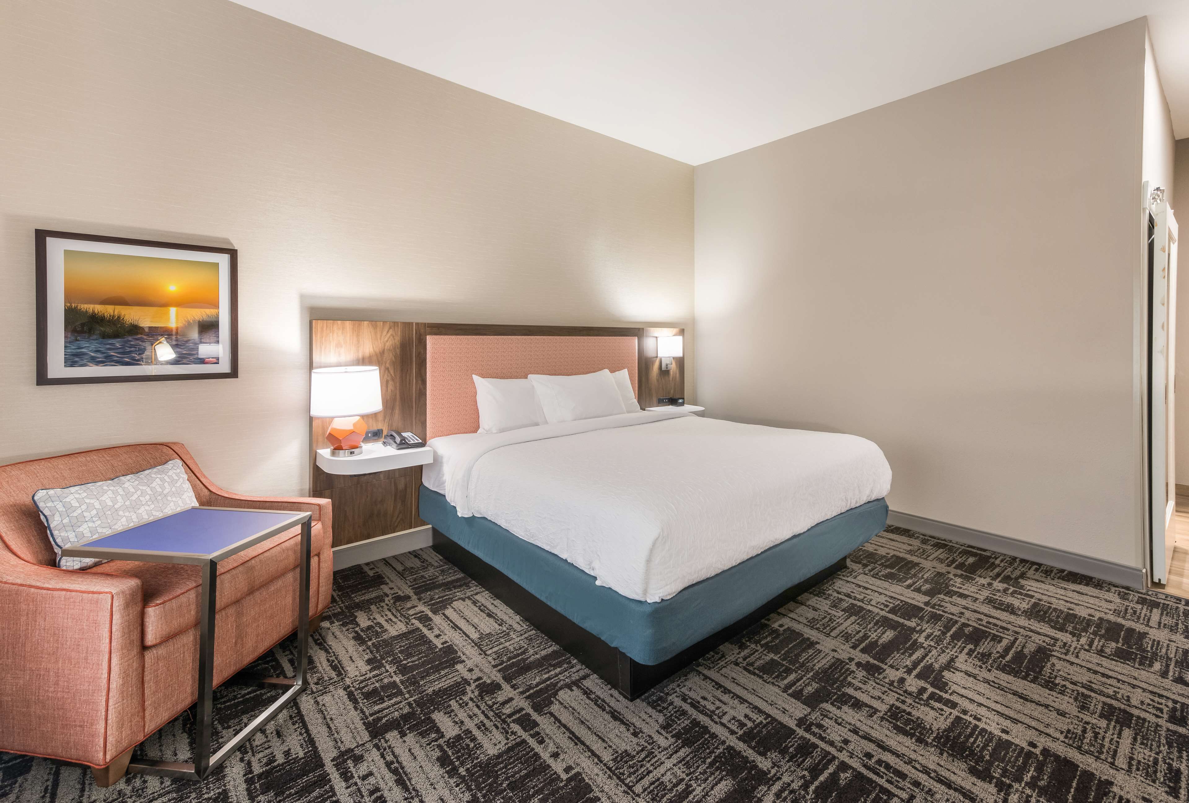 A hotel room with a bed, a desk, a chair, a nightstand, a lamp, and a framed picture on the wall.