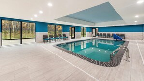 A modern indoor swimming pool with clear glass walls, a blue accent wall, and a seating area with chairs and a blue towel.