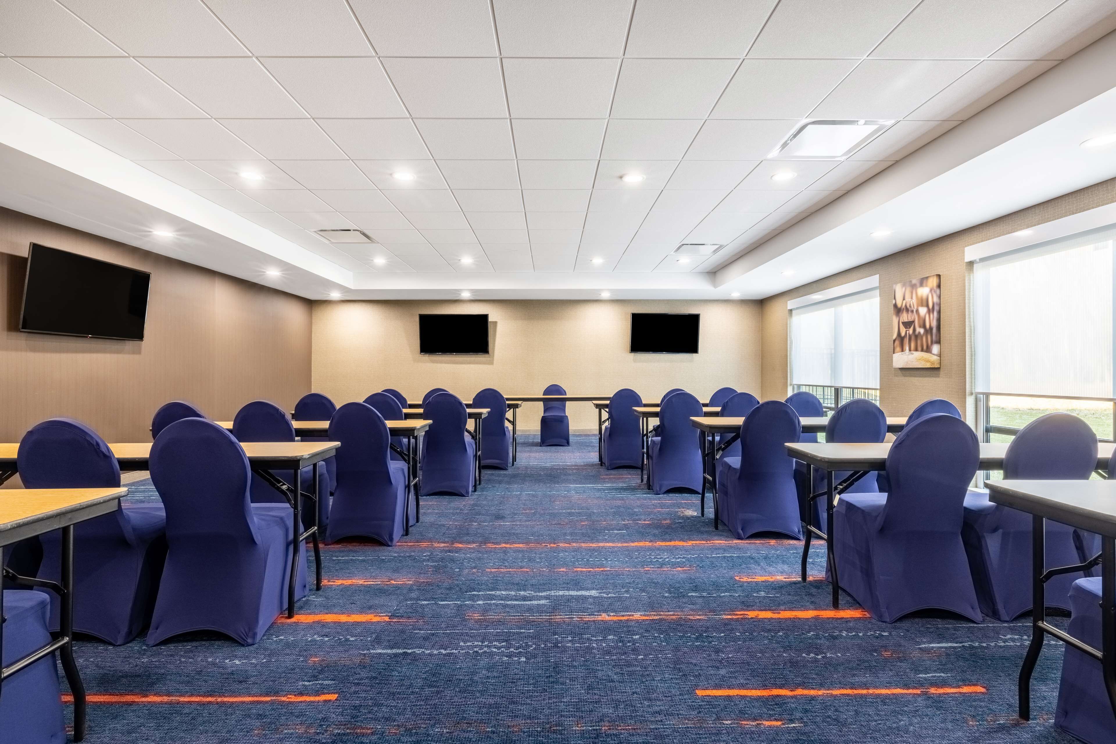 Un salón de conferencias con filas de mesas y sillas, una pantalla grande y piso alfombrado.