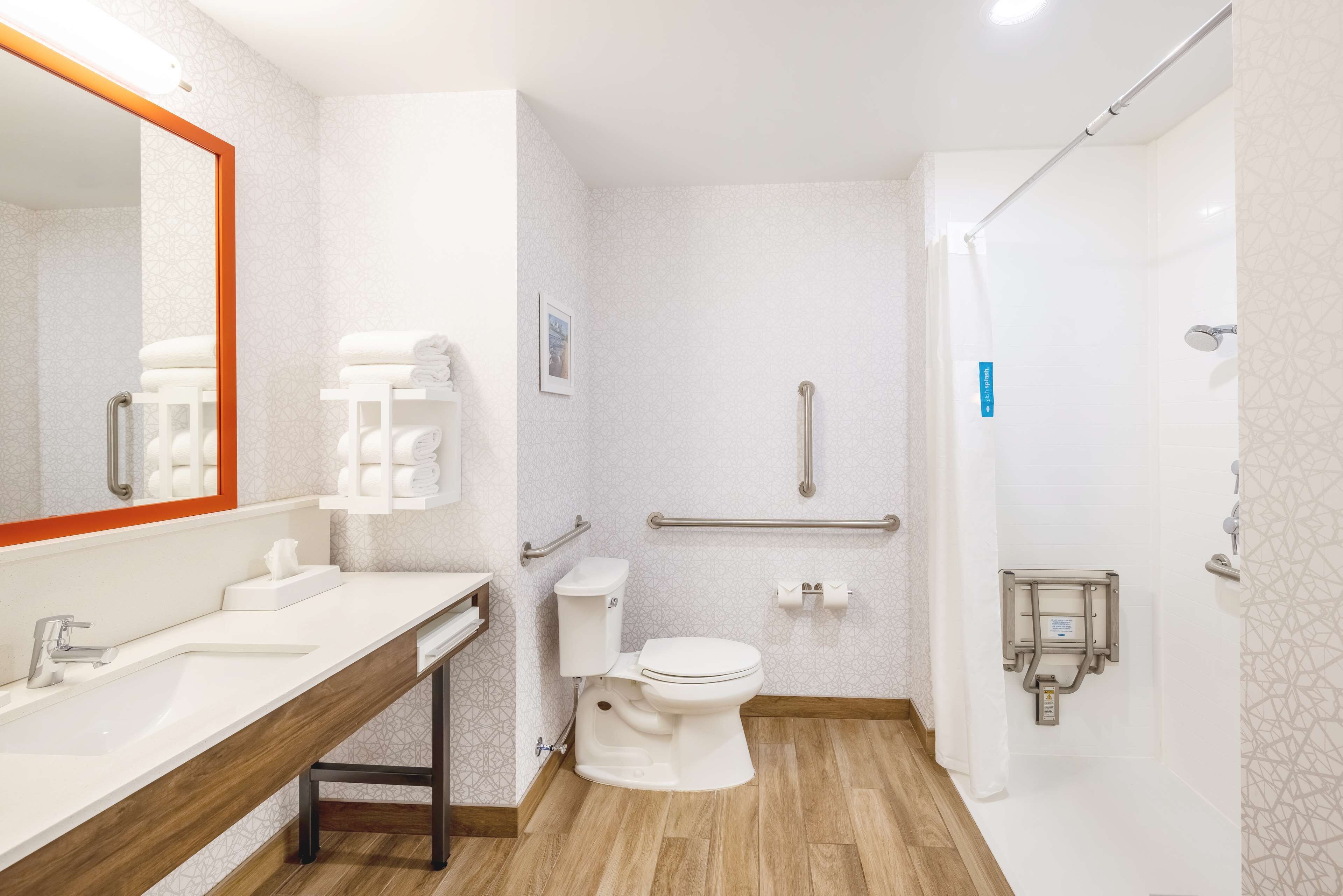 Un baño moderno con tocador de madera, inodoro blanco, ducha con barra de apoyo y dispensador de desinfectante de manos.