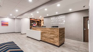 A reception area with a wooden reception desk, a welcome sign, a digital display, and a view of the hotel lobby.