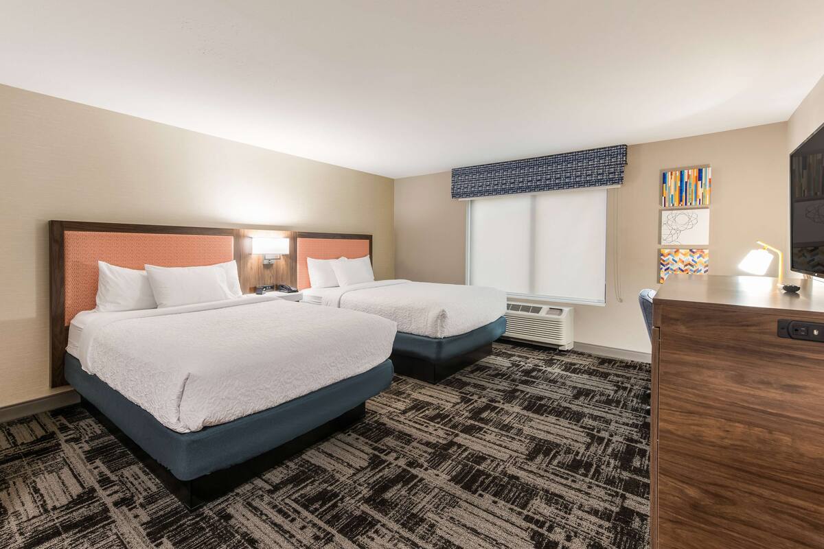 A hotel room with two beds, a desk, and a window with blinds.