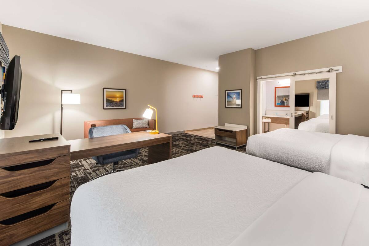 A hotel room with two beds, a desk with a lamp, a chair, a TV, and a mirror.