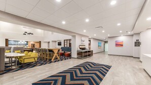 A spacious hotel lobby with a long zigzag patterned rug, a reception desk, and a seating area with yellow and gray furniture.