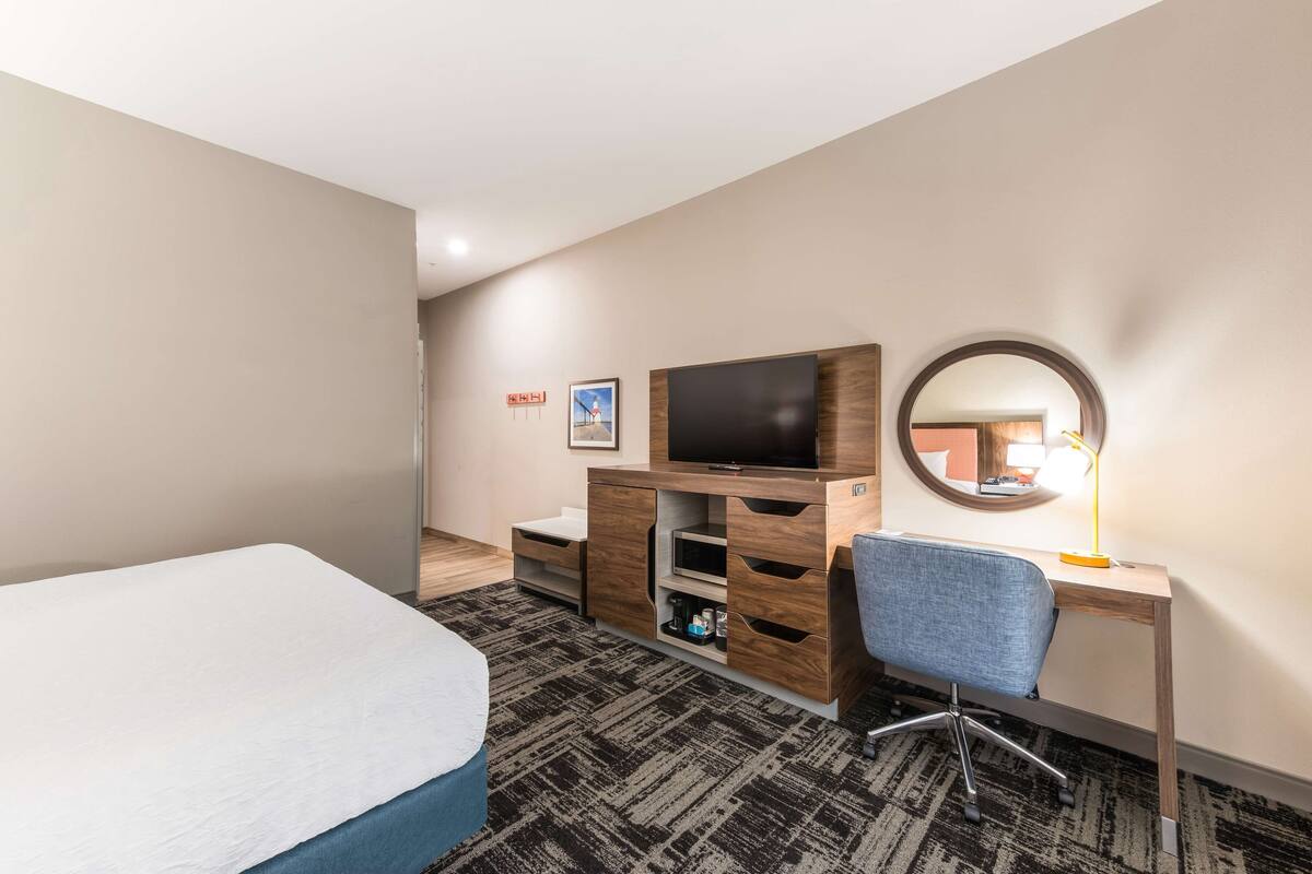 A hotel room with a bed, a desk with a chair, a TV, a mirror, and a carpeted floor.