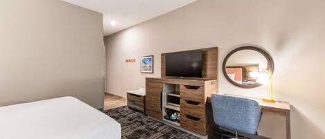 A hotel room with a bed, a desk with a chair, a TV, a mirror, and a carpeted floor.