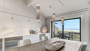 Un espace de salle à manger moderne, avec une table en bois, des chaises blanches et une vue sur l’océan grâce à de grandes fenêtres.