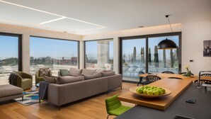 Una sala de estar moderna con un sofá, sillas, una mesa de comedor con manzanas y ventanas amplias que dan a vista de árboles y edificios.