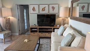 Un soggiorno con televisore, mobile TV in legno, divano beige, poltrona fantasia e tavolino con una conchiglia.