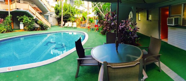 Une piscine avec une table et des chaises en verre, entourée de verdure et bordée d’un bâtiment doté d’un balcon.