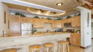 A kitchen with wooden cabinets, a stainless steel refrigerator, a microwave, and a granite countertop. There are three wooden stools at the counter. - Royal Waikiki Penthouses (Honolulu)