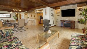 A spacious living area with a high ceiling, wooden beams, a glass coffee table, a sofa, a TV mounted on the wall, and a fireplace. - Royal Waikiki Penthouses (Honolulu)
