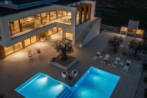 A modern multi-story house with a swimming pool, outdoor seating, and a well-lit patio area.