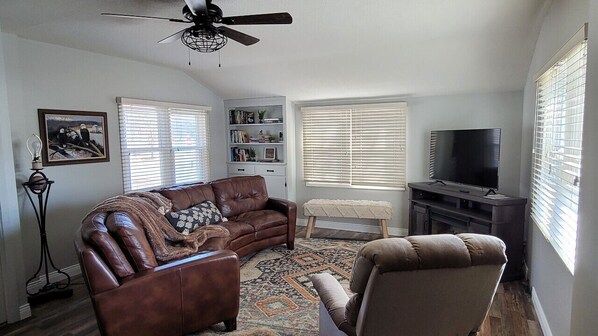 A living room with a brown leather sofa, a brown armchair, a bookshelf, a TV, and a ceiling fan.