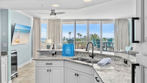 A modern kitchen with granite countertops, white cabinets, and a large window with a view of the ocean and palm trees.