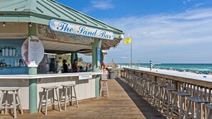 A beachside bar named "The Sand Bar" with a wooden deck, bar stools, and a view of the ocean.