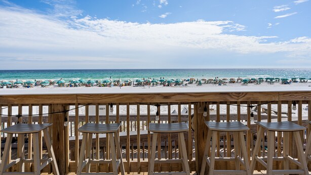 A wooden deck with bar stools overlooking a beach with umbrellas and people.