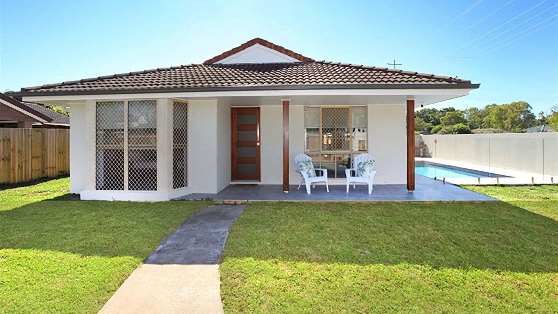 A single-story house with a tiled roof, a fenced backyard, a swimming pool, and a small patio with two chairs.